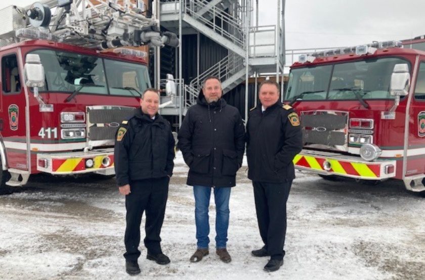 Une photo du maire en compagnie de deux membres du services incendie en face de deux camions de pompier.